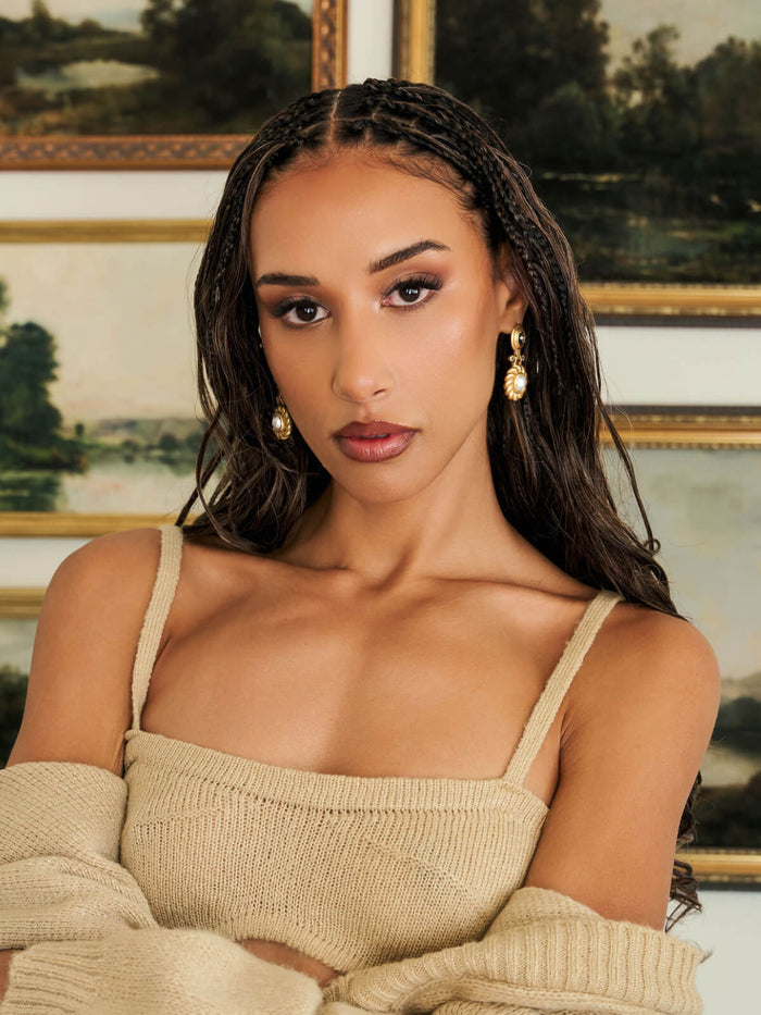 A woman with braided hair wearing a beige knit spaghetti-strap top and matching sweater draped off her shoulders, with gold earrings, posed in front of framed landscape paintings.