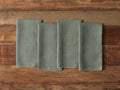 Four square linen napkins in a muted green color, with frayed edges, laid out side by side on a rustic wooden surface.