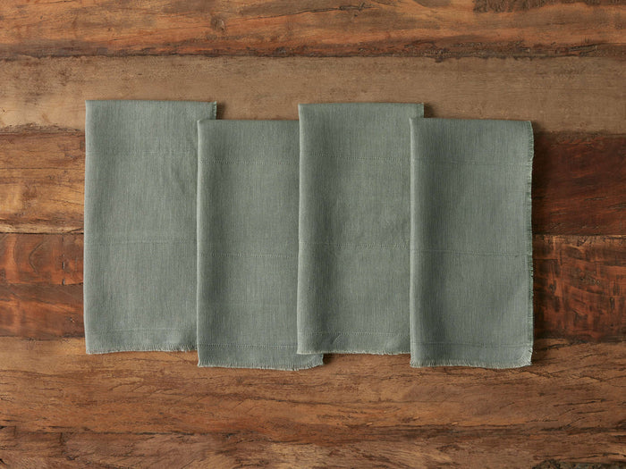 Four square linen napkins in a muted green color, with frayed edges, laid out side by side on a rustic wooden surface.