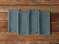 Four blue-gray linen napkins with frayed edges are neatly arranged on a rustic wooden table, highlighting their texture and casual style.