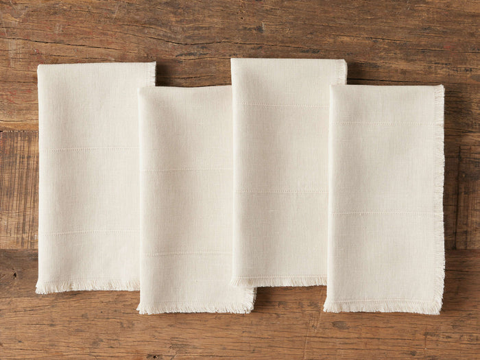 Set of four cream linen napkins arranged in a row on a wooden surface, featuring frayed edges and a textured handwoven pattern.