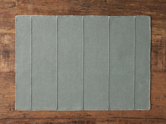 Linen Placemat in muted green with vertical stitched lines and frayed edges, laying flat on a wooden surface.