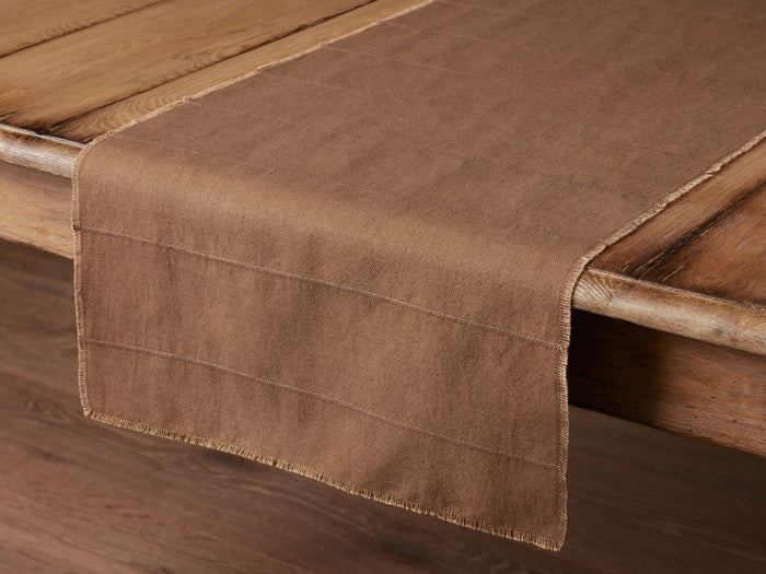 Linen Table Runner in brown, laid over a wooden table. Features frayed edges and handwoven texture for a rustic look.