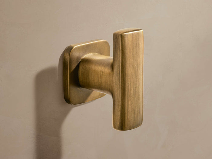 Logan Robe Hook in antique brass finish mounted on a beige wall, featuring a rectangular shape with rounded edges.