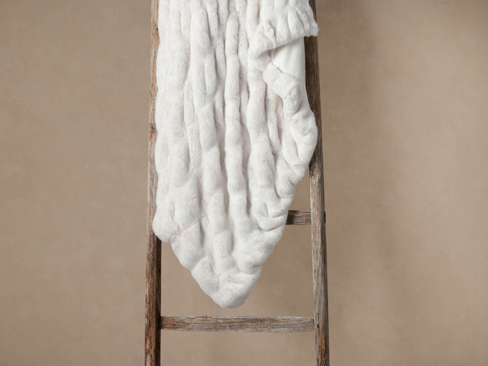 White Luxe Faux Fur Pleated Throw draped over a rustic wooden ladder, showing its soft, plush texture.