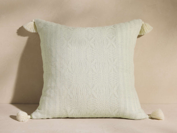 Madera Alpaca Tassel Pillow, light green with subtle woven pattern, featuring cream tassels on all four corners, on a neutral surface.