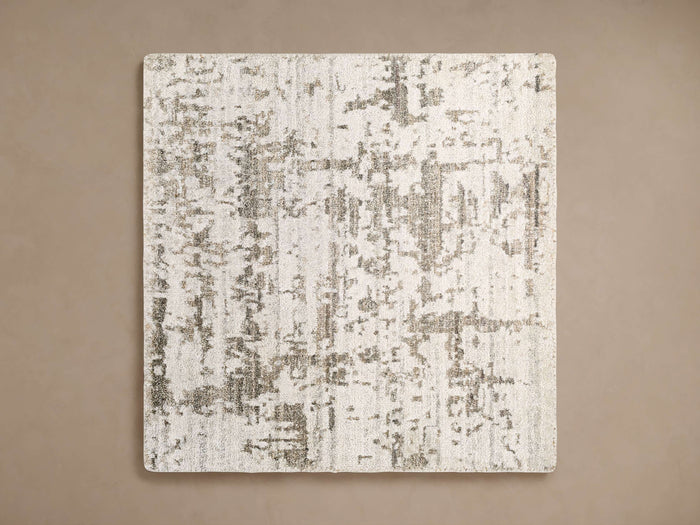 Madison Rug Swatch: rectangular fabric with an abstract, distressed pattern in off-white and brownish-grey, on a brown surface.