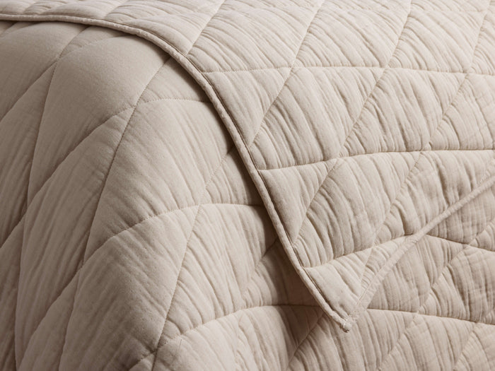 Close-up of a light beige Malea Cotton Quilt with a diamond quilted pattern, showing a folded edge and soft, crinkled texture.