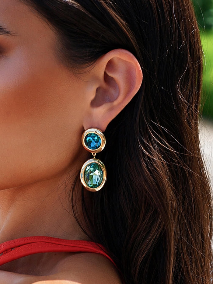 A close-up of a woman's ear wearing a gold drop earring with a round blue gemstone at the top and an oval green gemstone at the bottom.
