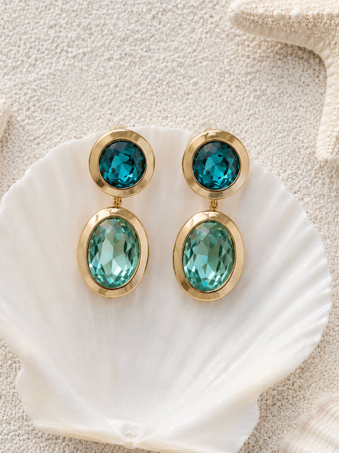 A pair of gold drop earrings with faceted teal blue gemstones, displayed on a white scallop shell over a textured beige background.