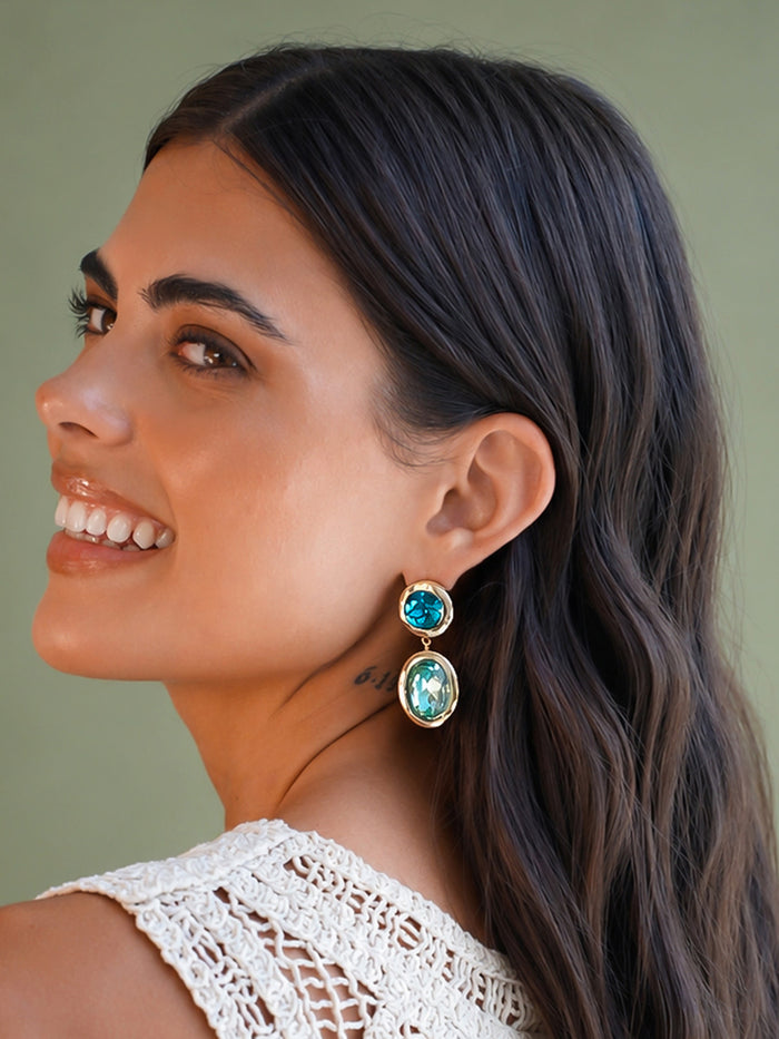 A close-up side profile of a woman with long dark hair wearing a gold drop earring with two oval turquoise-blue gemstones, set against a solid olive green background.