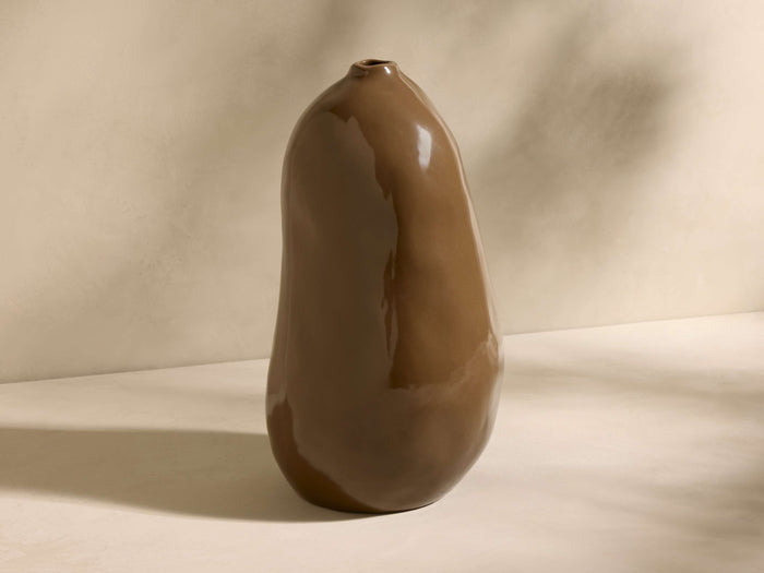 A glossy brown ceramic vase with a smooth, uneven surface, resembling a pear shape, placed on a light-toned surface.