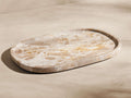 Mariyna Bath Tray: oval marble tray with natural beige, brown, and white veining, angled on a light background.