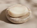 Mariyna Soap Dish: Round, beige marble with natural veining, holding an oval off-white soap bar, on a light neutral surface.