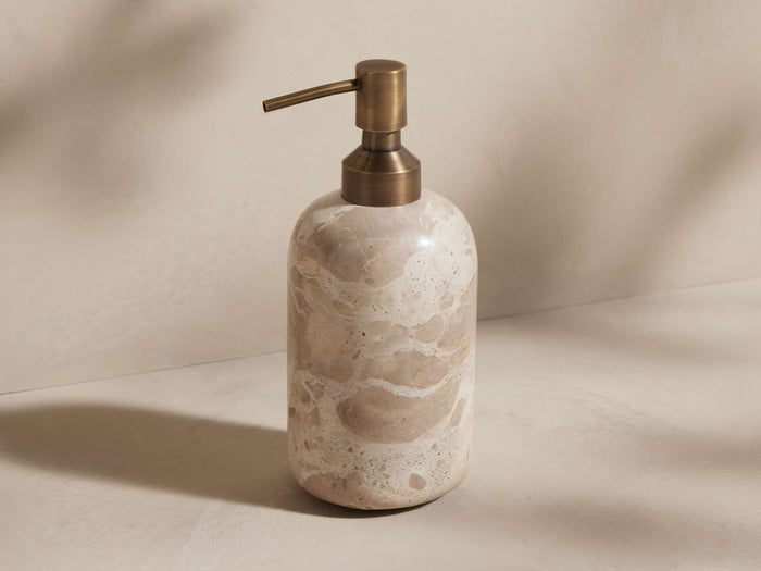 Mariyna Soap Pump. Round, beige marble dispenser with unique natural veining, topped by an antique brass pump, standing upright.