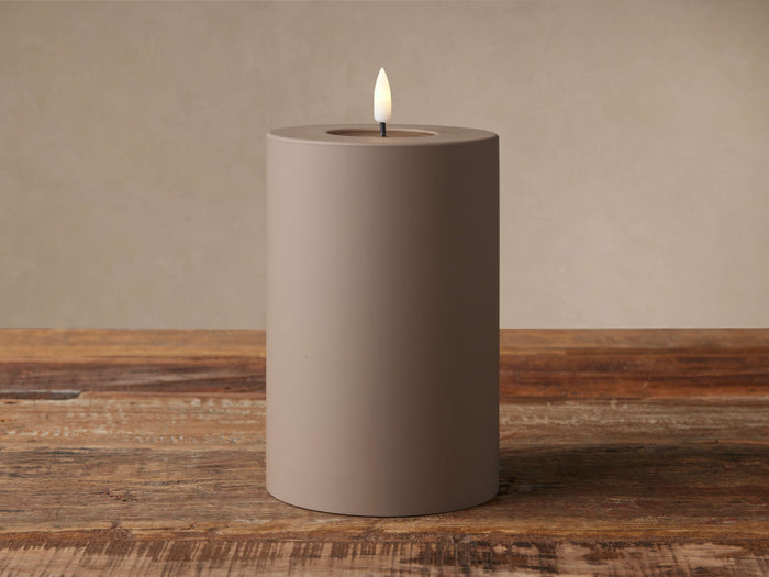 Matte brown cylindrical LED candle with a glowing faux flame, sitting on a rustic wooden surface against a beige wall.