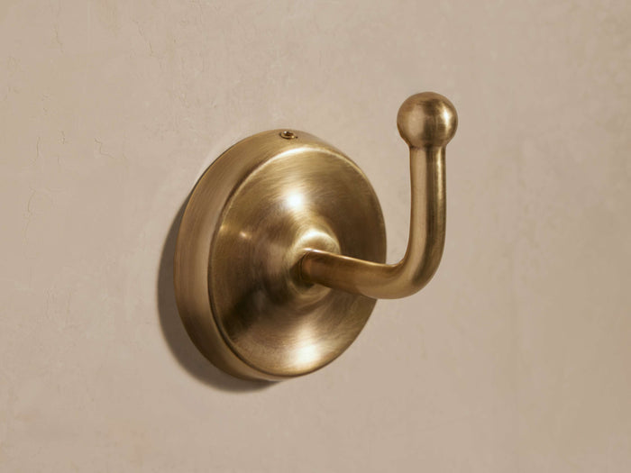 Maxwell Hook, antiqued brass, mounted on a light wall. Rounded base, curved arm with a spherical tip, facing right.