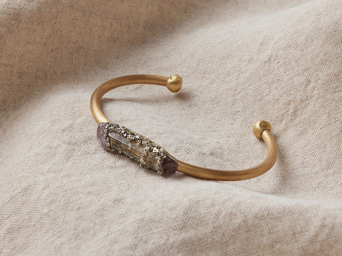Maylin Amethyst Cuff featuring a hand-shaped brass band with central amethyst crystal and pyrite accents, resting on fabric.