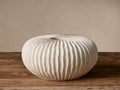 Meadow Vase, round, off-white ceramic with deeply ribbed texture, on a rustic wooden surface against a light wall.