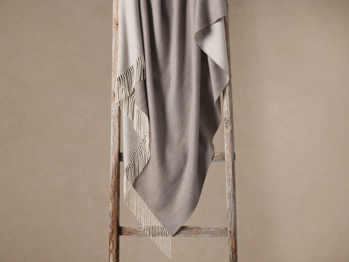 Melange Cashmere Throw draped on a rustic wooden ladder. The two-sided grey blanket features elegant white fringe details.