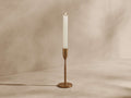Milo Taper Holder in antique gold with a textured finish, holding a lit white taper candle against a warm, textured wall.
