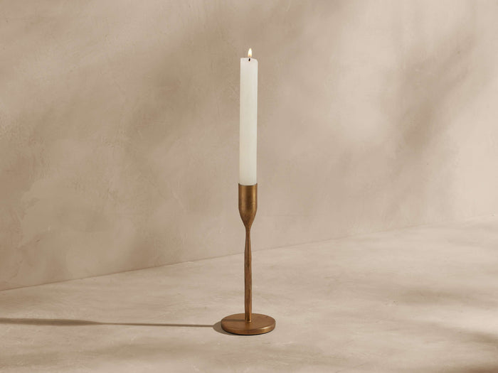 Milo Taper Holder in antique gold with a textured finish, holding a lit white taper candle against a warm, textured wall.