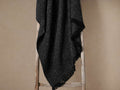 Montane Alpaca Throw in Onyx, dark grey, textured and fringed, draped casually over a rustic wooden ladder.