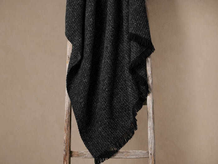 Montane Alpaca Throw in Onyx, dark grey, textured and fringed, draped casually over a rustic wooden ladder.