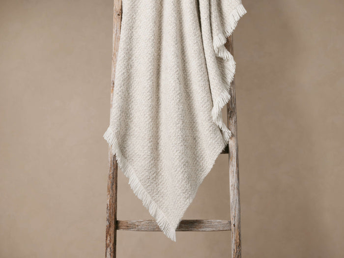 Montane Alpaca Throw, light beige with speckled texture and soft fringe, draped over a rustic wooden ladder against a wall.