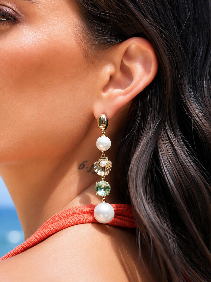 A close-up of a woman's ear wearing a long drop earring with a gold top, a white pearl bead, a gold scalloped shell charm, a green faceted bead, and a larger white pearl bead at the bottom.