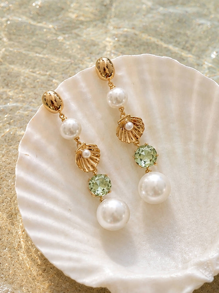 A pair of gold drop earrings with white pearls, gold shell charms, and light green faceted gemstones, displayed on a white seashell over a sandy beach background.