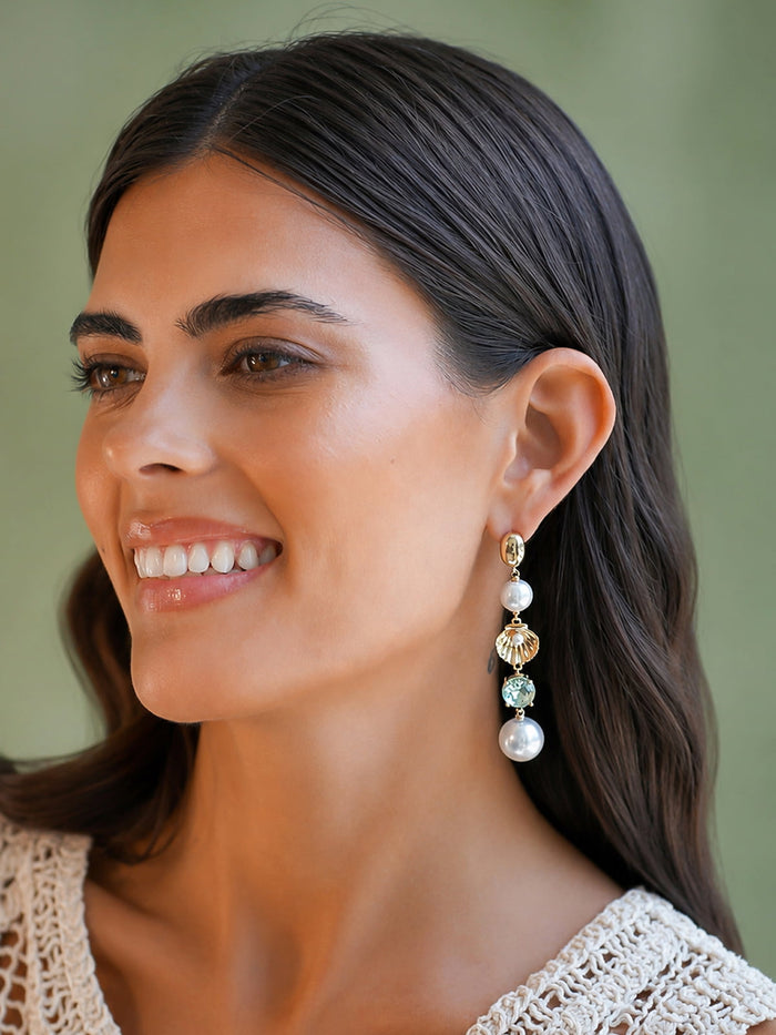 A close-up of a smiling woman with dark hair wearing a long drop earring with white pearl-like beads, a gold heart-shaped charm, and a light green faceted bead against a soft olive green background.