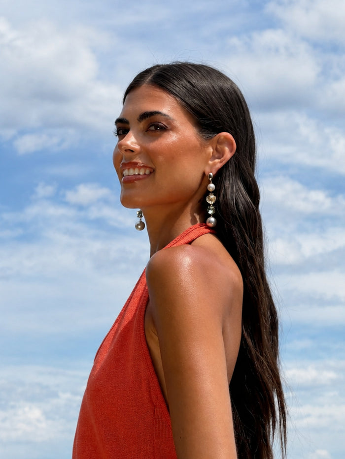 A woman with long dark hair is shown in profile outdoors against a blue sky with clouds, wearing a coral-red halter top and dangling pearl-like earrings.
