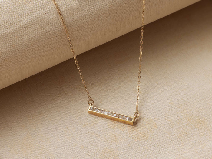 Morgana Gold Pendant Necklace with a horizontal rectangular gold bar set with small raw diamonds on a delicate chain, placed on fabric.
