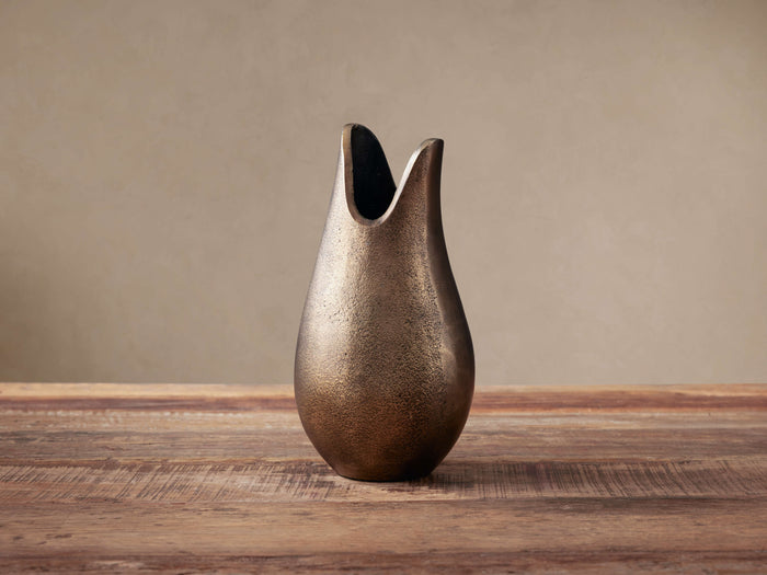 Myles Bud Vase: organic, sculptural aluminum vase with antiqued brass finish and unique flowing top, on a rustic wooden table.