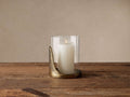 Myles Hurricane: A clear glass cloche atop an organic antiqued brass base, holding a lit white pillar candle on wood.