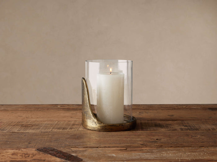 Myles Hurricane: A clear glass cloche atop an organic antiqued brass base, holding a lit white pillar candle on wood.