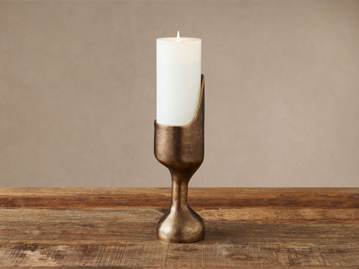 Myles Pillar Holder, antiqued brass cast aluminum, organically shaped, holds a lit white pillar candle on a wood surface.