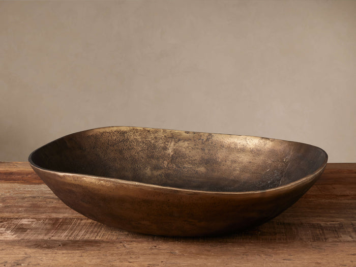 Myles Round Bowl made of cast aluminum with a textured antiqued brass finish, sitting on a rustic wooden table against a neutral wall.