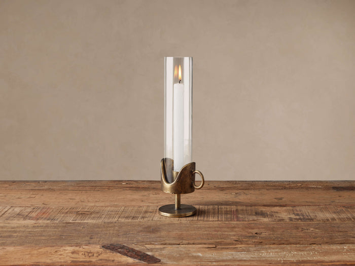 Myles Taper Holder with an antiqued brass sculptural base, clear glass cloche, and a lit white taper candle on a wooden table.