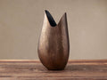 Sculptural Myles Vase in antiqued brass, textured, with a peaked opening, standing on a rustic wooden table against a plain wall.