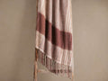 Soft, fringed throw blanket draped over a rustic wooden ladder. Features warm brown stripes on a light beige background, with long tassels.