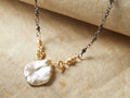 Nadya Necklace featuring a lustrous, irregularly-shaped pendant with gold accents on a delicate chain, displayed on fabric.
