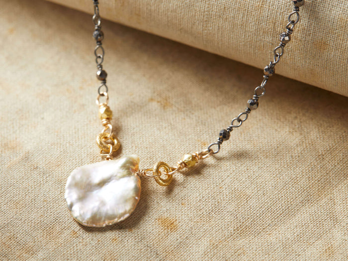 Nadya Necklace featuring a lustrous, irregularly-shaped pendant with gold accents on a delicate chain, displayed on fabric.