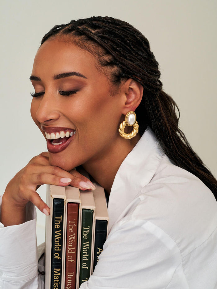A woman with braided hair wearing a white shirt and large gold hoop earrings with white pearl centers, smiling and leaning against a stack of books.
