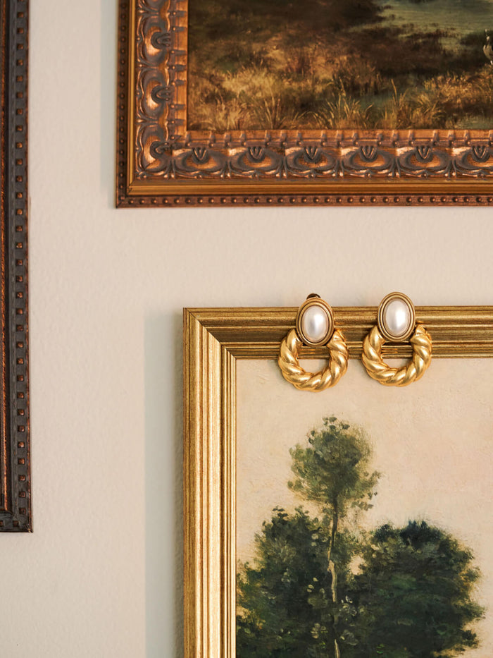 A pair of gold-toned hoop earrings with oval pearl-like centers, displayed on the corner of a gold picture frame against a light beige wall.