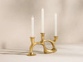 Nouria 3-Light Taper Holder: Golden, organic brass candelabra with three white taper candles, two lit, on a light surface.