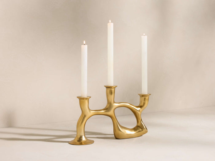 Nouria 3-Light Taper Holder: Golden, organic brass candelabra with three white taper candles, two lit, on a light surface.
