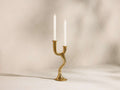 Nouria Double Taper Holder: golden, sculptural wavy design, holding two lit white taper candles on a light surface.