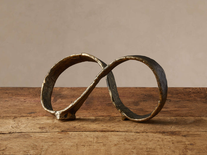 Nowles Sculpture, an antique brass distressed aluminum infinity symbol, sits on a rustic wooden table against a light wall.